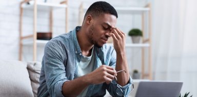 A Black man holding glasses in one hand, and pressing his fingers from his other hand into his forehead. He looks tired.