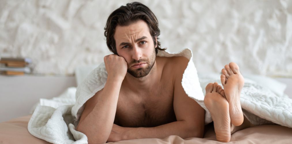 A man laying in bed looking disappointed. A woman's feet are near his head.