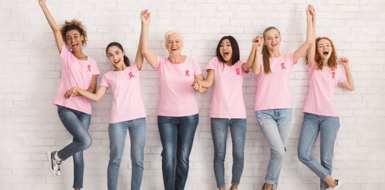 A group of women in pink shirts with breast cancer ribbons pinned to their chests. They are holding hands and cheering.