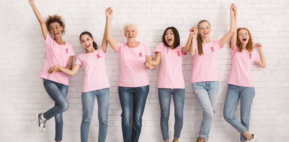 A group of women in pink shirts with breast cancer ribbons pinned to their chests. They are holding hands and cheering.